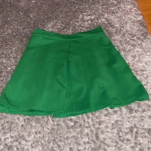 Grön kortkjol från FB Sister - Snygg grön kortkjol från FB Sister i storlek M. Kjolen har en A-linjeformad siluett och är tillverkad i ett mjukt polyestertyg. Den stängs med en diskret dragkedja bak och har en enkel, stilren design utan mönster eller extra detaljer.