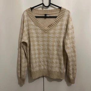 Beige rutig stickad tröja H&M - Säljer en beige stickad tröja från H&M med v-ringad hals och rutigt mönster i ljusbeige och vitt. Tröjan har långa ärmar och ribbade muddar vid hals, ärmslut och nederkant. Perfekt för dig som gillar chill och snygg stil.