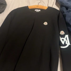 Svart sweatshirt från Moncler - Svart sweatshirt från Moncler med klassisk rund halsringning. Tröjan har Moncler-logga på bröstet och ärmen samt en vit grafisk detalj på ena ärmen. Perfekt för dig som gillar stilrena och exklusiva plagg. Storlek XXL men sitter som XL 