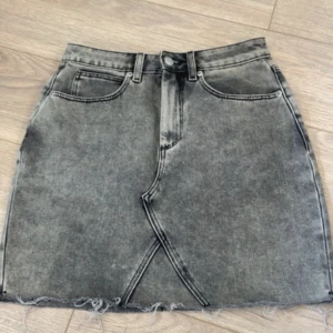 Grå jeanskjol med rå kant - Säljer en grå jeanskjol med cool tvättad look och rå, fransig kant nedtill. Kjolen har klassiska fickor fram och bak, bälteshällor och en snygg omlott-detalj framtill. Perfekt för dig som gillar streetstyle och vill ha en edgy vibe.