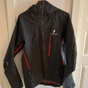 Svart vindjacka Peak Performance M - Peak Performance svart vindjacka med röda dragkedjor och huva. Mycket fint skick, 