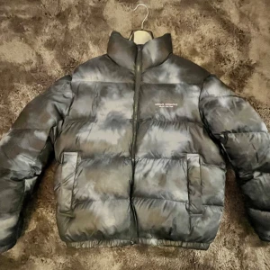 Svart pufferjacka från Armani Exchange - Säljer en fet svart pufferjacka från Armani Exchange med hög krage och diskret logga på bröstet. Jackan har dragkedja framtill, två sidofickor och en cool mörkgrå tonad effekt. Perfekt för kalla dagar och riktigt snygg streetstil.