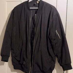 Svart bomberjacka med dragkedjor - Svart bomberjacka från H&M med klassisk ribbad krage, muddar och nederkant. Jackan har silvriga dragkedjor framtill och på ärmen samt två snedställda fickor. Oversized passform som ger en cool och avslappnad vibe.