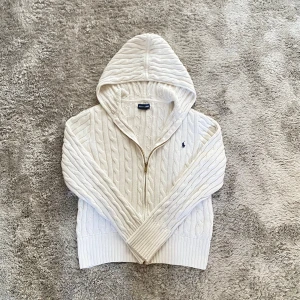 Vit kabelstickad kofta från Ralph Lauren - Superfin vit kabelstickad hoodie från Ralph Lauren med dragkedja och broderad logga på bröstet. Tröjan är i 100% bomull, har långa ärmar, ribbade muddar och en mysig huva. Perfekt för lager-på-lager och chill dagar.