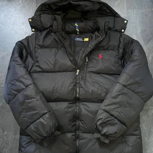 Svart pufferjacka från Polo Ralph Lauren - Svart pufferjacka från Polo Ralph Lauren med röd broderad logga på bröstet. Jackan har huva, tvåvägsdragkedja och elastiska muddar vid ärmslut. Materialet är syntet och jackan är quiltad för extra värme. Perfekt för kalla dagar.
