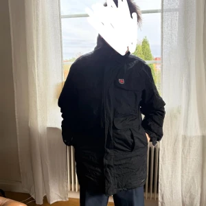 Svart parkajacka från Fjällräven - Svart parkajacka från Fjällräven med klassisk röd logga på bröstet. Jackan har flera stora fickor framtill, hög krage och dragkedja med vindslå. Materialet är slitstark polyester och jackan har en loose passform, perfekt för kalla dagar.dragkedjan fungerar men själv kroken eller va det kallas har gått av! 