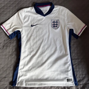 England Nike vit fotbollströja - England fotbollströja från Nike i vitt med marinblå detaljer och krage. Tröjan har korta ärmar med röd och blå rand på mudden, broderad Nike-logga och England-emblem på bröstet. Tillverkad i lätt och ventilerande polyester.
