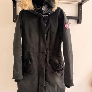 Svart parkas från Canada Goose - Svart parkas från Canada Goose med päls på huvan och klassisk logga på ärmen. Jackan har långa ärmar, två stora fickor framtill och dragkedja med knappslå. Perfekt för kalla vinterdagar och har en snygg, stilren look. Såklart äkta.