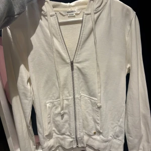 Vit hoodie från Zadig & Voltaire med paljetter - Säljer en vit hoodie från Zadig & Voltaire med dragkedja, huva och snörning. På ryggen står det 'ROCK' i guldiga paljetter med blixtdetalj. Tröjan har långa ärmar och två fickor framtill. Perfekt för dig som gillar coola detaljer. 