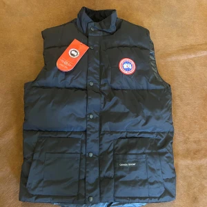 Svart dunväst från Canada Goose - Svart dunväst från Canada Goose med klassisk logga på bröstet, två stora fickor framtill och tryckknappar längs hela fronten. Västen har hög krage och diskret dragkedja i sidan. Perfekt för kalla dagar och har en clean, stilren look.