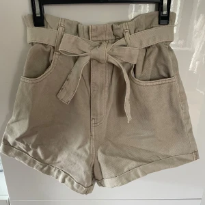 Beige highwaist shorts med knytband - Snygga beige shorts med hög midja och brett knytband i midjan. Modellen har stora fickor fram och bak samt uppvikta benslut. Perfekta för varma dagar och ger en chill men trendig vibe.