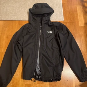Svart regnjacka från The North Face - Svart regnjacka från The North Face med huva och meshfoder. Jackan har dragkedja framtill, snörning i nederkant och logga på både bröstet och ryggen. Jackan är knappt använd.
