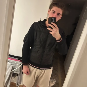 Svart Moncler bomberjacka med huva - Svart bomberjacka från Moncler med huva och coola detaljer som ficka med dragkedja på ärmen. Jackan har ribbade muddar med röd, vit och blå rand, samt Moncler-logga på ärmen. Materialmix av polyester och bomull, perfekt för dig som gillar streetwear.