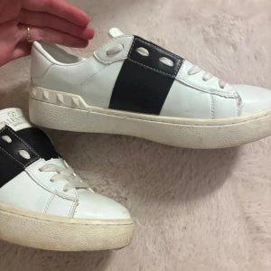 Valentino Garavani vita skor - Snygga vita sneakers från Valentino Garavani med bred svart läderdetalj över snörningen och ikoniska pyramidnitar på hälen. Skorna har rund tå, platt sula och vita snören. Perfekt för dig som gillar exklusiv och stilren design.