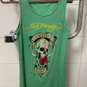 Ed Hardy linne / topp - PRIS KAN DISKUTERAS VID SNABB AFFÄR! Går för ca 600-1500kr på andra sidor - Säljer en grön linnetopp från Ed Hardy med ikoniskt döskalletryck, röda rosor och texten 'Love Dies Hard'. Toppen har gul Ed Hardy-logga framtill och baktill, är ärmlös och ribbad i materialet. Vintage, säljs inte längre så är väldigt exklusiv. Stretchig så passar allt från S-L. Passar mig med S men även min vän som är en L