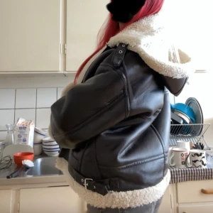 Svart teddyjacka med fuskpäls - Säljer en svart teddyjacka med fluffig vit fuskpäls på krage, ärmslut och nederkant. Jackan har en loose passform, dragkedja framtill och snygga spänndetaljer på sidorna. Perfekt för dig som vill ha en varm och trendig look under höst och vinter.