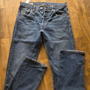 Levi's 501 blå jeans, straight fit W28 L32 - Klassiska Levi's 501 jeans i blå denim med raka ben och knappgylf. Snygga kontrastsömmar och ikonisk läderpatch bak i midjan. Modellen har fem fickor och den röda Levi's-taggen på bakfickan. Perfekt för dig som gillar tidlös stil.
