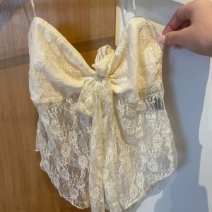 Krämvit spets tubtopp från SHEIN - Supersöt krämvit tubtopp från SHEIN i spets med stora rosettdetaljer framtill. Toppen är axelbandslös och har ett transparent, blommigt spetsmönster som ger en romantisk vibe. Perfekt för dig som vill sticka ut med en feminin och trendig look.