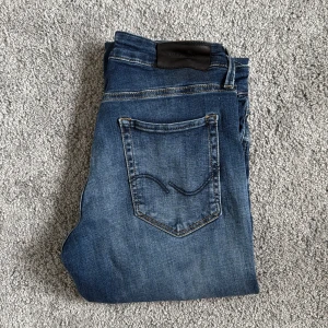 Blå slim jeans från Jack & Jones - Snygga blå jeans från Jack & Jones med klassisk fyraficksdesign och slim passform. Jeansen har kontrastsömmar, knapp och dragkedja i gylfen samt en mörk tvätt med lätt slitning. Perfekta för dig som gillar en smal siluett och stilren look.