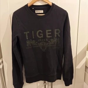 Svart sweatshirt från Tiger of Sweden - Svart sweatshirt från Tiger of Sweden Jeans med tryck framtill i samma ton. Klassisk rund halsringning och långa ärmar. Mjuk insida och ribbade muddar vid ärmslut och nederkant. Perfekt för en avslappnad och stilren look.