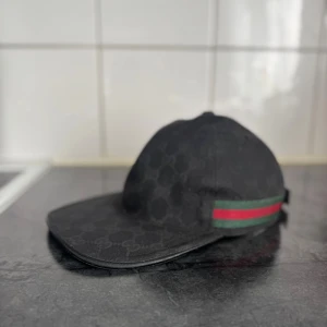 Svart keps från Gucci med logomönster - Säljer en svart keps från Gucci med diskret GG-logomönster över hela kepsen. Klassisk böjd skärm och justerbar rem bak i skinn. Sidan har den ikoniska gröna och röda randen. Tillverkad i Italien och i bomullsmaterial med detaljer i skinn.