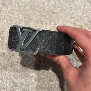 Bälte - Säljer ett svart bälte med ikoniskt spänne i silverfärg. Bältet har diskret monogrammönster och är gjort i skinn med ljus insida. Perfekt accessoar för att lyfta din outfit med en lyxig touch.