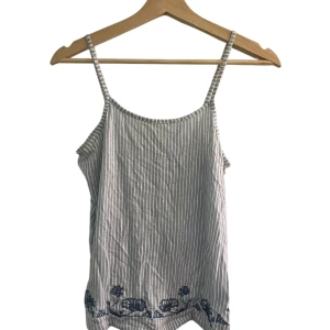 Randig linnetopp med broderi - Snygg randig linnetopp med tunna axelband och blå broderade blommor längst ner. Toppen är vit med smala blå ränder och har en avslappnad passform, perfekt för varma dagar.