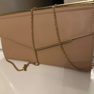 Beige kuvertväska med guldkedja - Snygg beige kuvertväska med stilren design och guldfärgade detaljer. Väskan har en elegant guldkedja som axelrem och ett dekorativt guldfärgat metallband på framsidan. Perfekt för att lyfta din outfit med en chic touch.