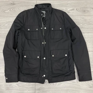 Svart fieldjacka från J.Lindeberg - Stilren svart jacka från J.Lindeberg med fyra stora fickor framtill och quiltat foder. Jackan har hög krage, dragkedja och tryckknappar. Perfekt för dig som vill ha en snygg och funktionell jacka med klassisk look. Ända negativa med jackan är att kedjan hoppar ur ibland därav priset!