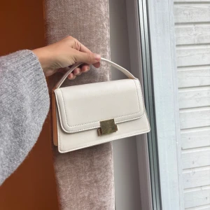 Vit handväska med gulddetalj - Stilren vit handväska i skinnimitation med rektangulär form och elegant guldfärgat spänne framtill. Väskan har ett kort handtag och minimalistisk design som ger en clean och modern look.