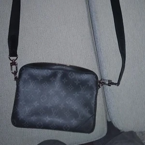Svart axelväska Louis Vuitton - Snygg svart axelväska från Louis Vuitton med klassiskt monogrammönster. Väskan har justerbar axelrem, dragkedja upptill och är tillverkad i skinn. Perfekt storlek för det viktigaste och en clean look som funkar till många outfits. 