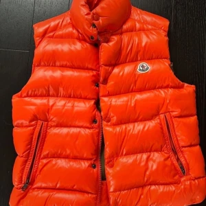 Orange dunväst från Moncler - Snygg orange dunväst från Moncler med hög krage och två dragkedjefickor framtill. Nyskick, självklart äkta. Storlek 2=M