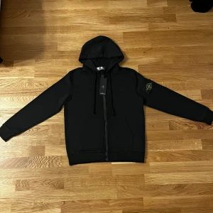 Svart hoodie från Stone Island - Svart hoodiejacka från Stone Island med klassisk logotyp-patch på vänster ärm. Jackan har huva med snörning, hel dragkedja framtill och två fickor. Materialet är mjukt och känns som bomull, perfekt för en chill och stilren look.
