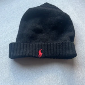 Svart stickad mössa Polo Ralph Lauren - Svart stickad mössa från Polo Ralph Lauren med klassisk röd logobroderi framtill. Mössan har ribbad kant och är tillverkad i mjukt material som håller dig varm. Perfekt accessoar för kyliga dagar och snygg till streetwear.