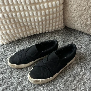 Svarta slip-on skor från Anna Field - Svarta slip-on skor från Anna Field med twistad detalj över tån och vit platt sula. Skorna är tillverkade i bomullstyg och har en rund tå. Insidan är dekorerad med små vita prickar för en extra touch. Perfekta för dig som gillar enkel och stilren design.