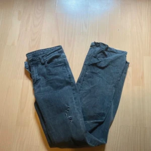 Mörkgrå bootcut jeans med slitna detaljer - Säljer ett par mörkgrå jeans med coola slitningar vid knäet. De har några skavanker, en längst ner där de blivit slitna och sen har det gått sönder där man har bältet igenom men inget som märks. 
