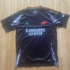 Arsenal svart bortatröja Adidas XL - Snygg svart Arsenal bortatröja från Adidas med gröna och röda detaljer på krage och axlar. Tryck med Emirates Fly Better på bröstet och klubbmärke i rött. Tillverkad i lätt och ventilerande polyester, perfekt för match eller träning.