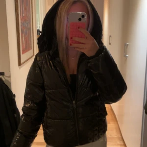 Svart glansig vinterjacka från Nelly - Säljer denna populära vinterjacka från Nelly! Original priset är 800kr. Svart glansig pufferjacka med huva och dragkedja framtill. Perfekt nu till vintern. En del av fickan har en liten lagning som inte syns.💞