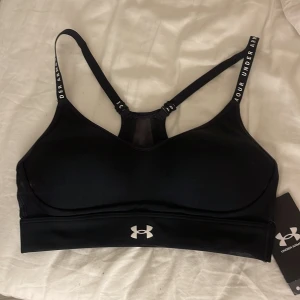Svart sporttopp från Under Armour XS - Sportig svart linnetopp från Under Armour med smala axelband och logga framtill. Toppen har meshdetalj i ryggen och justerbara band med text. Perfekt för träning och har en tight passform som sitter skönt mot kroppen.