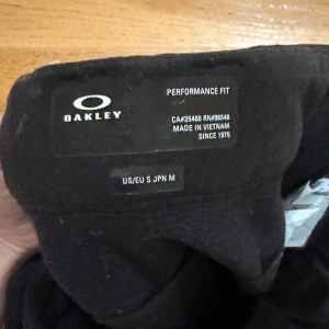 Svarta täckbyxor från Oakley, S/M - Svarta täckbyxor från Oakley med performance fit. Byxorna har två framfickor med dragkedja, bälteshällor och stilren design. Materialet är slitstarkt och passar perfekt för utomhusaktiviteter. Snyggt Oakley-logo på benet.
