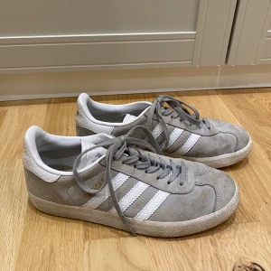 Adidas Gazelle grå/vita sneakers - Säljer ett par Adidas Gazelle sneakers i grå mocka med vita detaljer och klassiska tre ränder i präglat mönster. Skorna har snörning, vit sula och Adidas-logga bak. Perfekta för dig som gillar stilrena och tidlösa sneakers. Hård knut på ena snöret som ej går upp (se bild) men syns inte när man knutit dem