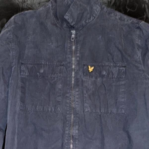 Mörkblå overshirt från Lyle & Scott - Snygg mörkblå overshirt från Lyle & Scott med dragkedja framtill och två bröstfickor. Jackan har klassisk ståkrage och den ikoniska gula loggan på vänster bröstficka. Perfekt för lager-på-lager och stilren look.