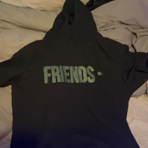 Svart Vlone Friends- hoodie med orm - Svart hoodie från Vlone med stor 'FRIENDS-' text på bröstet och ikoniskt V-tryck med röd och vit orm på ryggen. Klassisk huva och lång ärm, perfekt streetwear-plagg för dig som gillar unika detaljer och hypekultur.