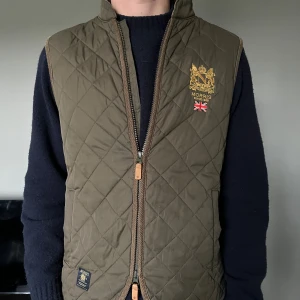 Olivgrön quiltad väst från Morris - Quiltad olivgrön väst från Morris med guldfärgat broderi och brittisk flagga på bröstet. Västen har ståkrage, dragkedja framtill och diskreta fickor. Perfekt för lager-på-lager och snygg till jeans eller chinos.