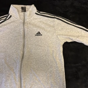 Grå Adidas fleecejacka med dragkedja - Snygg grå fleecejacka från Adidas med klassiska svarta ränder längs axlar och ärmar. Jackan har hel dragkedja framtill, hög krage och Adidas-logga på bröstet. Insidan är mjuk och varm fleece, perfekt för chill eller träning. Långärmad och sportig stil.