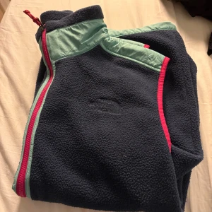 Blå fleeceväst från The North Face - Snygg blå fleeceväst från The North Face med ljusblå quiltade detaljer och två stora fickor framtill. Västen har en hög krage, rosa dragkedja och kontrastkant i ljusblått. Perfekt för lager-på-lager och chill dagar utomhus.