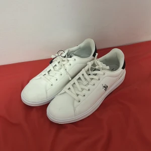 Vita sneakers från U.S. Polo Assn. - Snygga vita sneakers från U.S. Polo Assn. med svart detalj på hälen och logga på sidan. Skorna har snörning, rund tå och platt sula. Materialet är syntet med textilfoder. Röd och marinblå rand på baksidan ger en sportig vibe. Perfekta för dig som gillar stilrena och klassiska sneakers.