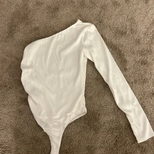 Vit oneshoulder topp från NA-KD - Säljer en stilren vit oneshoulder topp från NA-KD i slätt, glansigt material. Toppen har lång ärm på ena sidan och är av bodysuit-modell, vilket ger en snygg och tight passform. Perfekt för dig som vill sticka ut med en asymmetrisk look. Använd ett fåtal gånger 