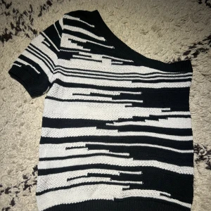 Svartvit stickad offshoulder tröja - Snygg stickad tröja med svart och vit randigt mönster i ett coolt glitch-inspirerat design. Tröjan har offshoulder och korta ärmar, vilket ger en trendig vibe. Perfekt för dig som gillar att sticka ut med din stil och vill ha något unikt i garderoben.