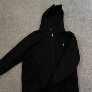 Svart hoodie från Polo Ralph Lauren - En svart Ralph Lauren zip hoodie med vit ikon. Mycket bra kvalitet och ett mycket bra kvalitets pris! Matchar med 1000+ outfits. M med passar som S
